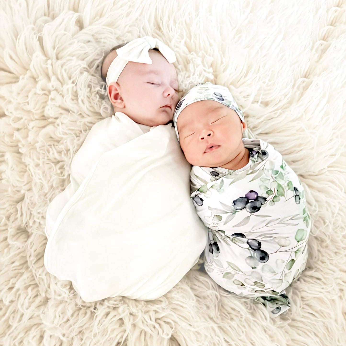 Two newborn babies swaddled in white and patterned wraps on a fluffy beige surface.