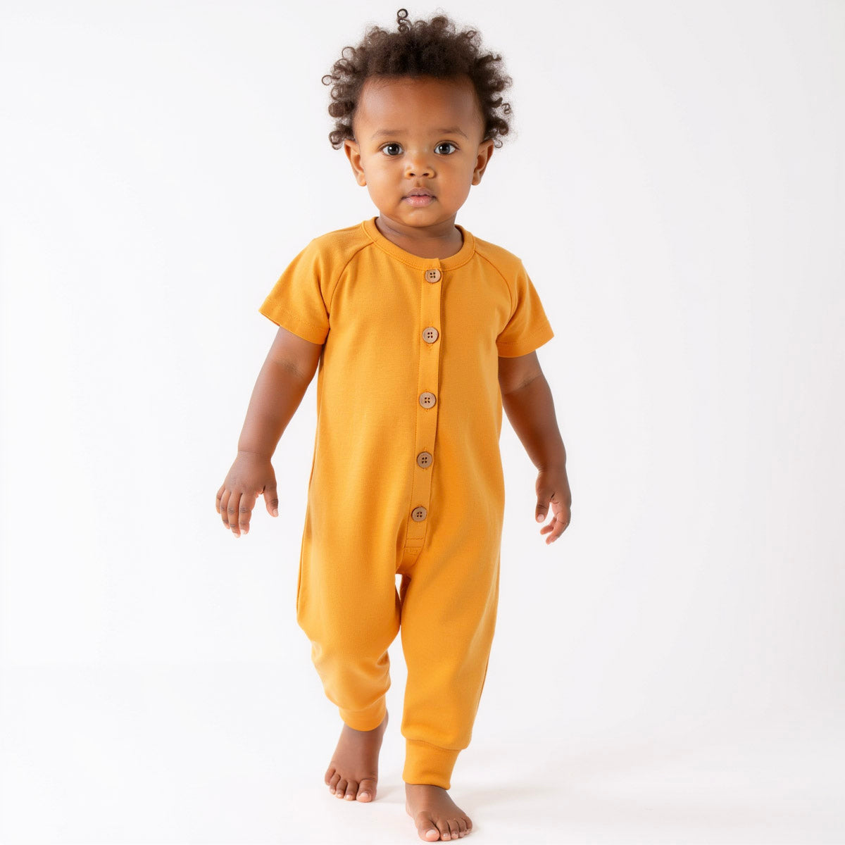A toddler wearing an orange short-sleeve romper with wooden button front closure, standing against a white background.