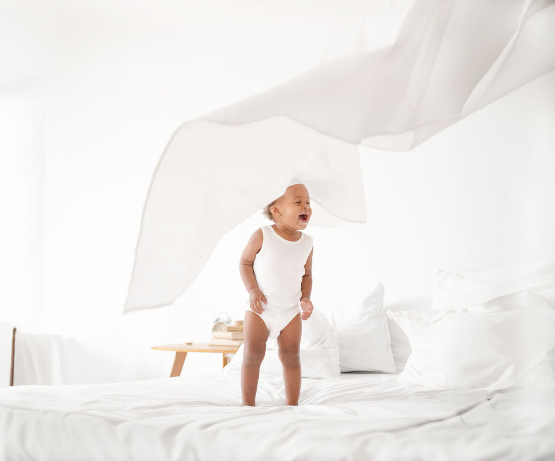 Toddler standing up on a bed wearing white clothing. 