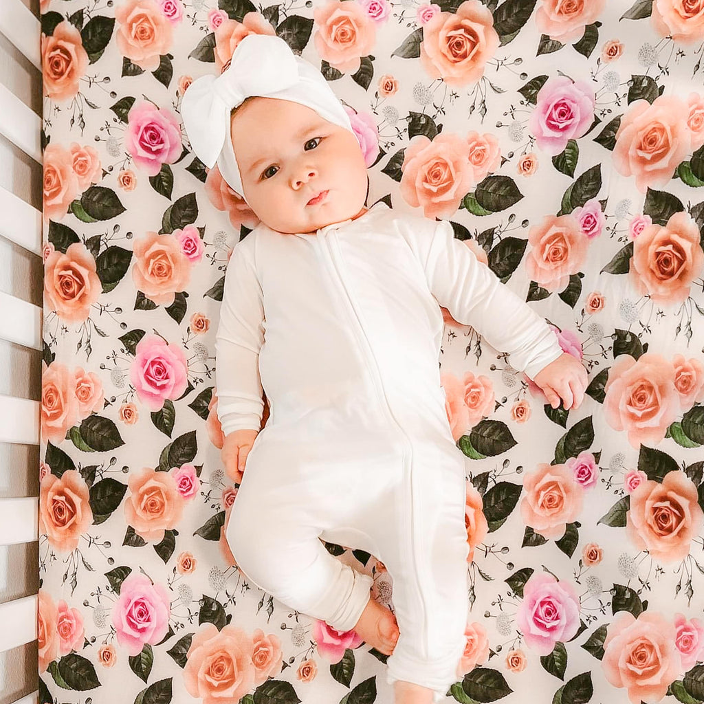Baby girl wearing white baby and toddler one piece pajamas while lying down on a floral crib sheet 