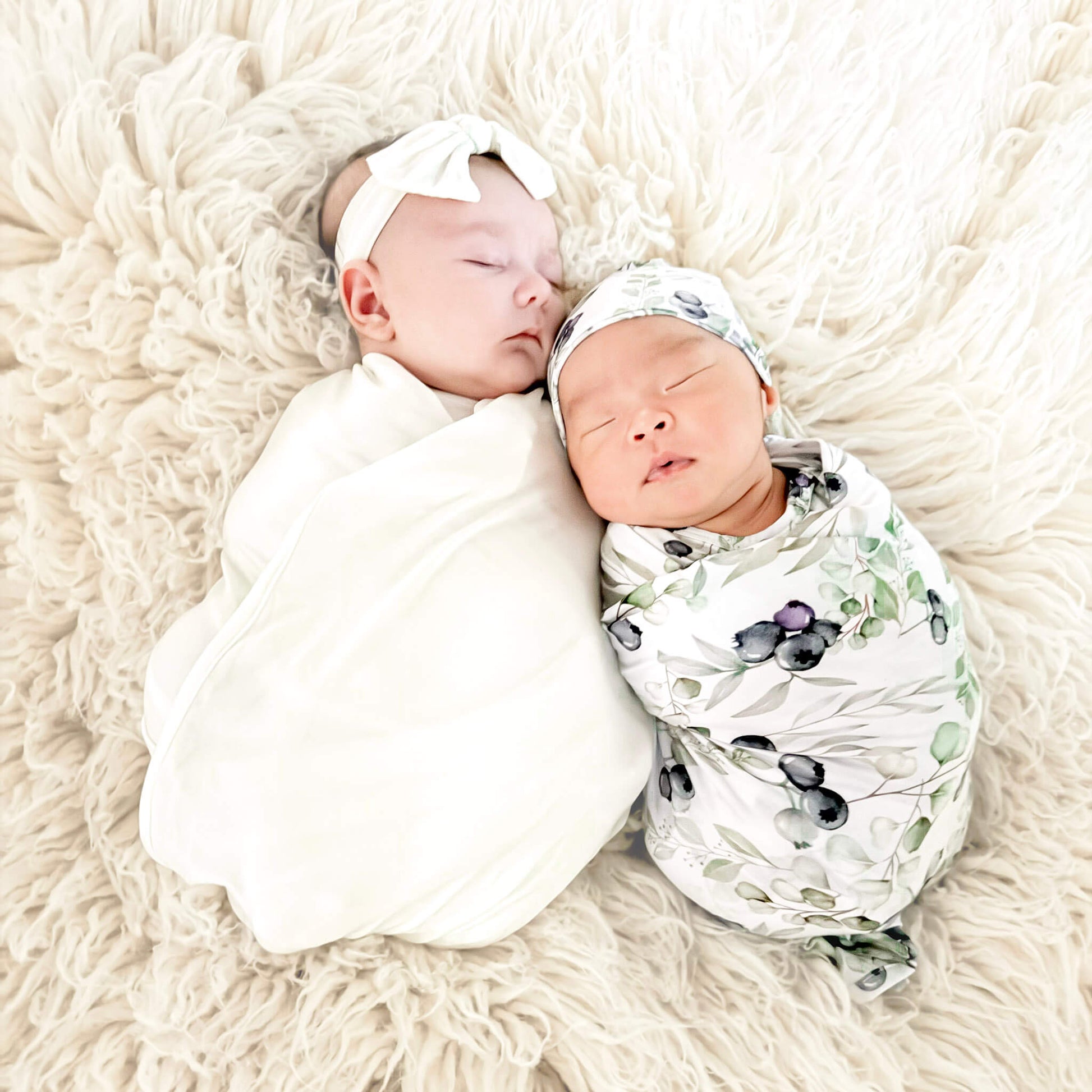 Two newborn babies swaddled in white and patterned wraps on a fluffy beige surface.