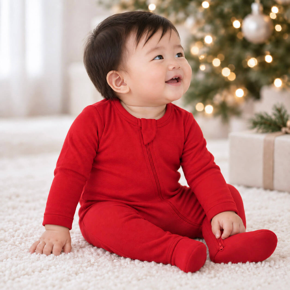 A baby sitting on a white carpet in front of a softly blurred Christmas tree and gift box, wearing a bright red long-sleeve footed one-piece with a front zipper.