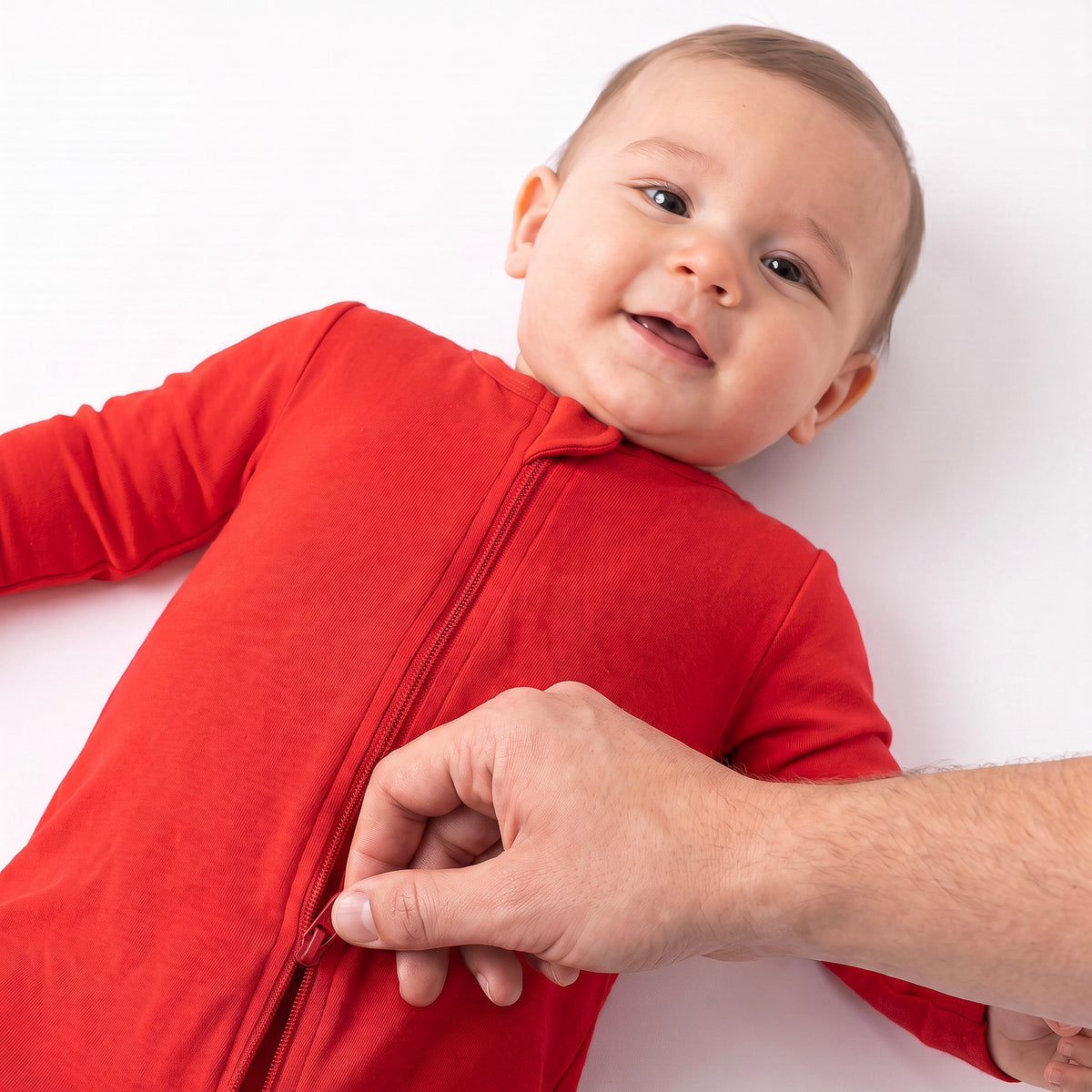 A baby wearing a bright red zip-up sleeper lies on a white background while an adult hand helps pull the zipper open.