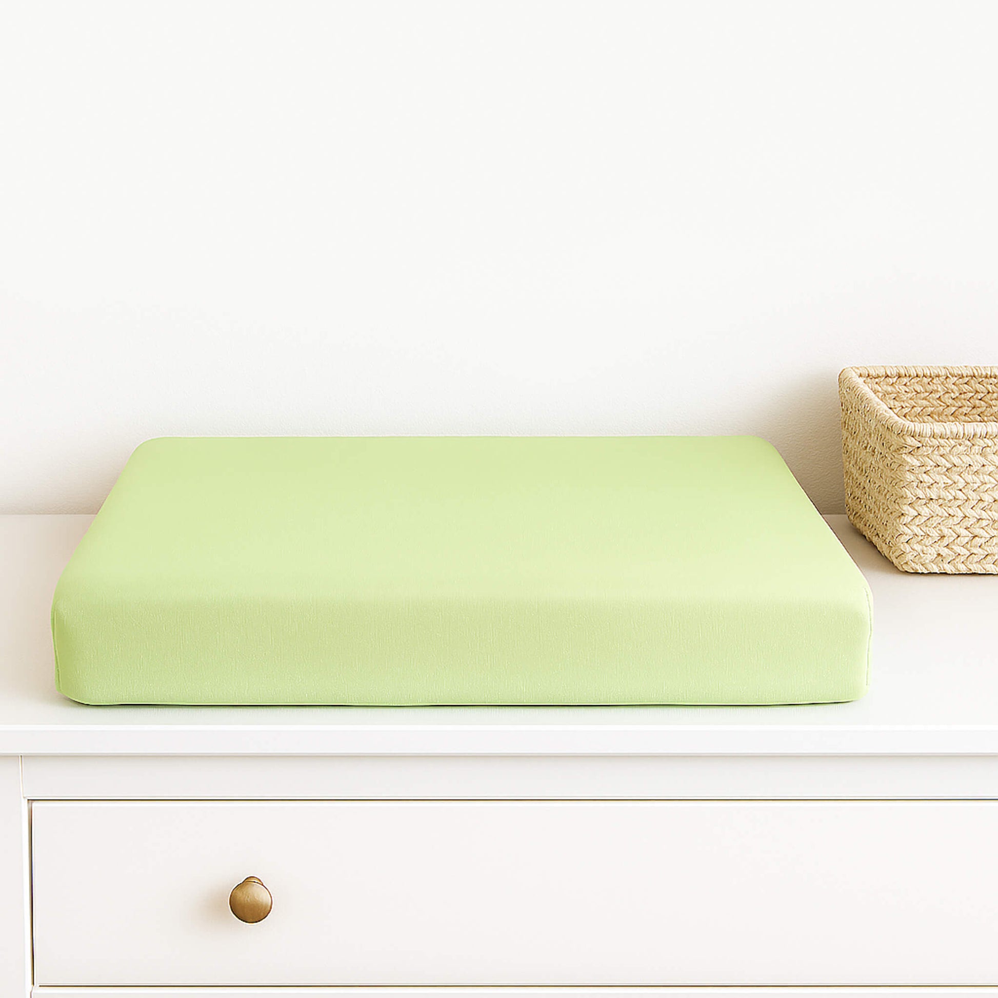 Light green changing pad cover on a white dresser with a woven basket in the background