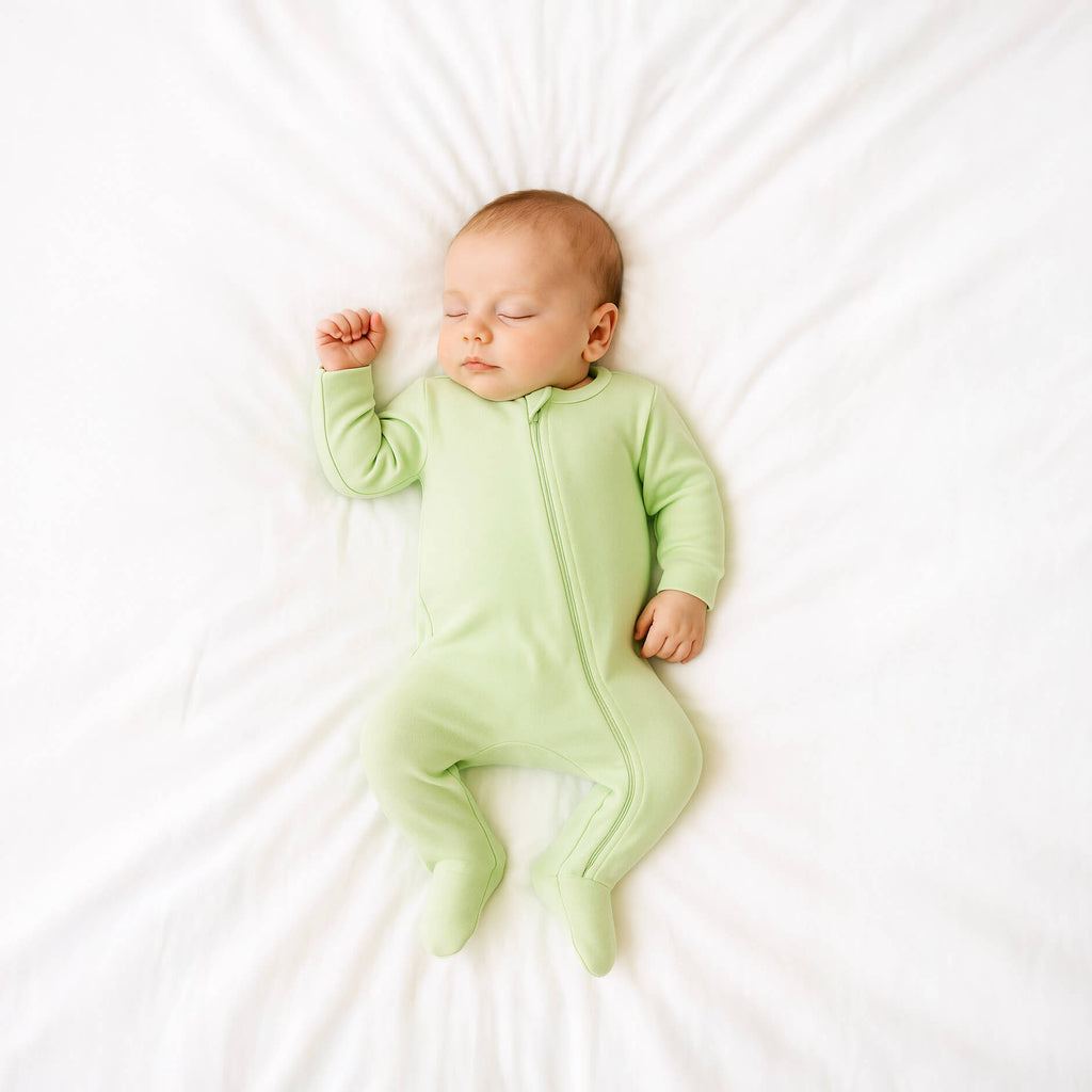 Baby in a green onesie sleeping on a white surface