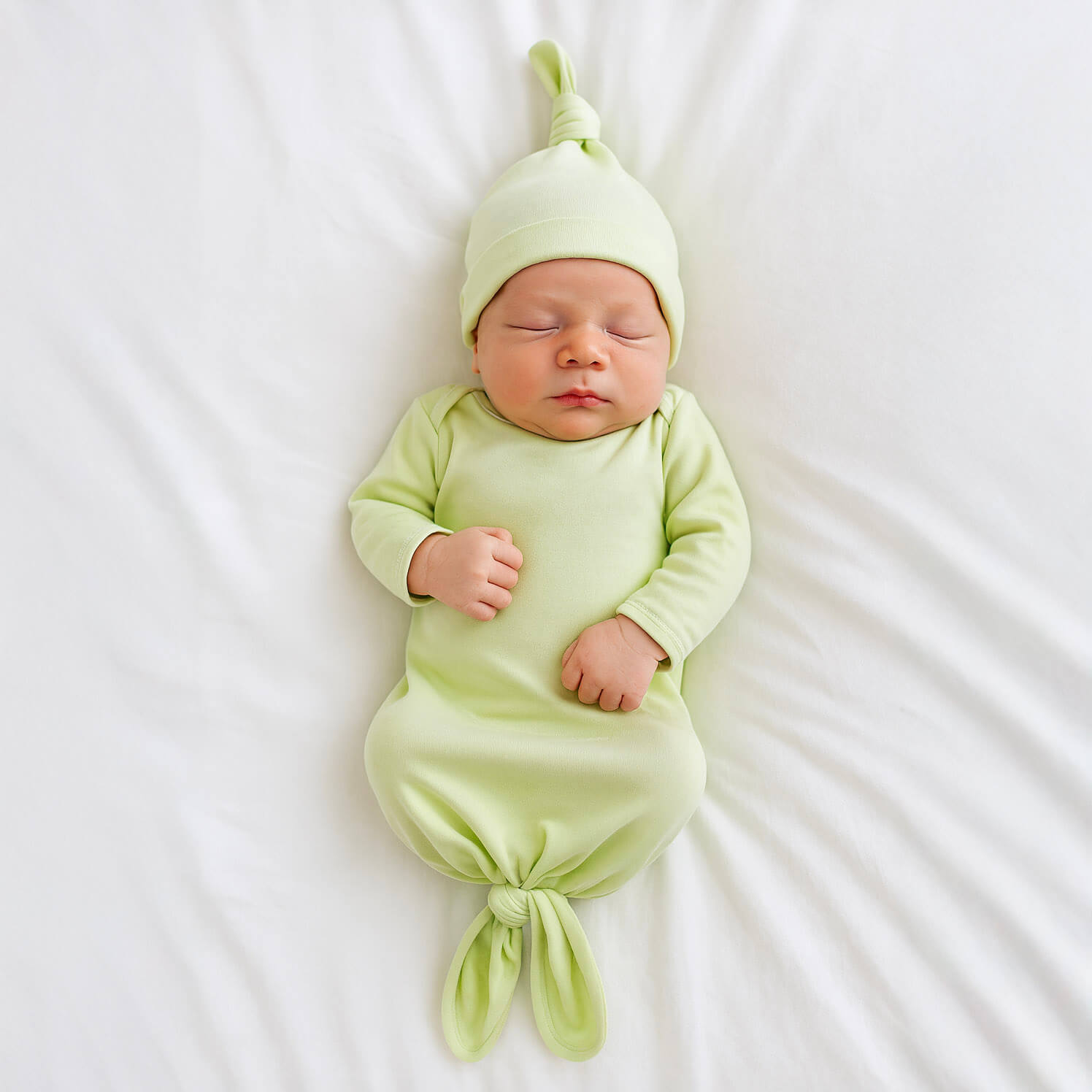 Newborn baby in a light green gown and hat lying on a white surface