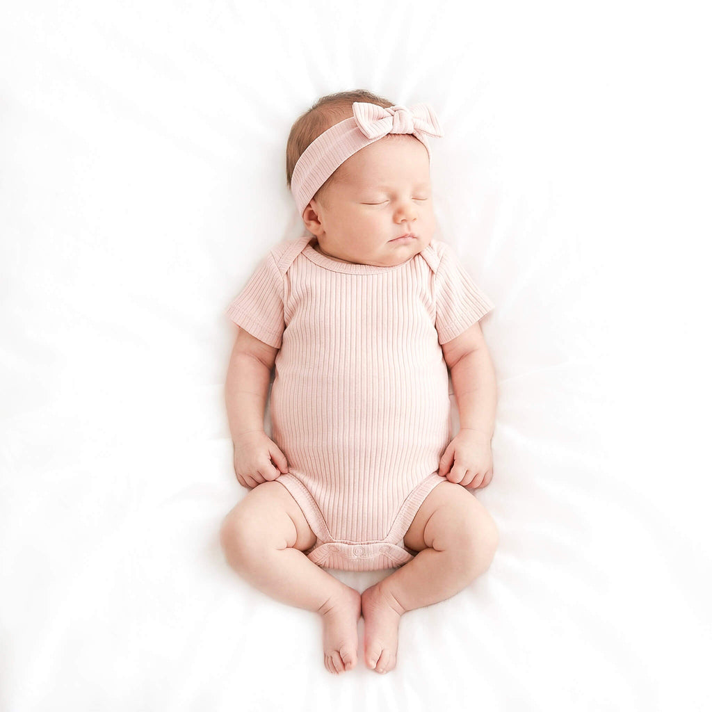 Newborn baby wearing a pink ribbed romper and headband on a white background