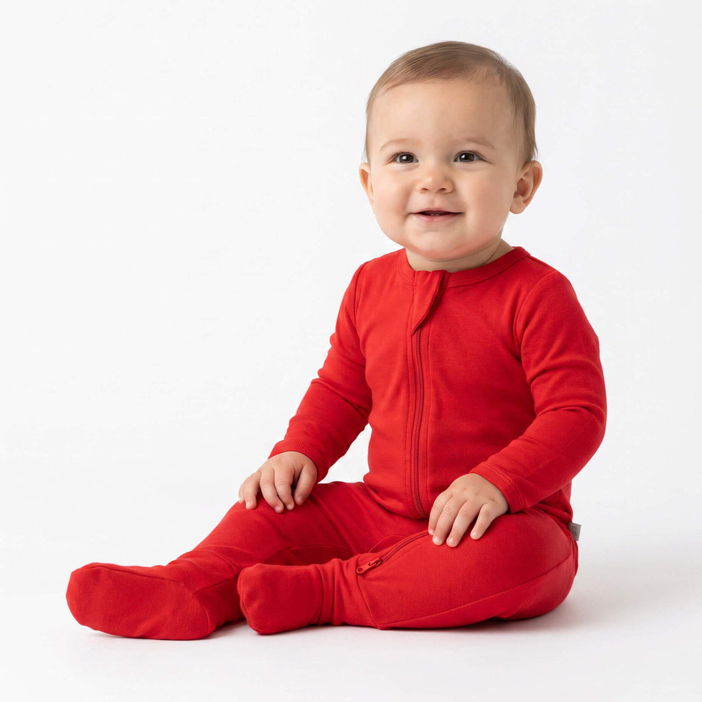 A smiling baby sitting cross-legged on a clean white studio background, wearing a bright red long-sleeve zip-front footed one-piece.