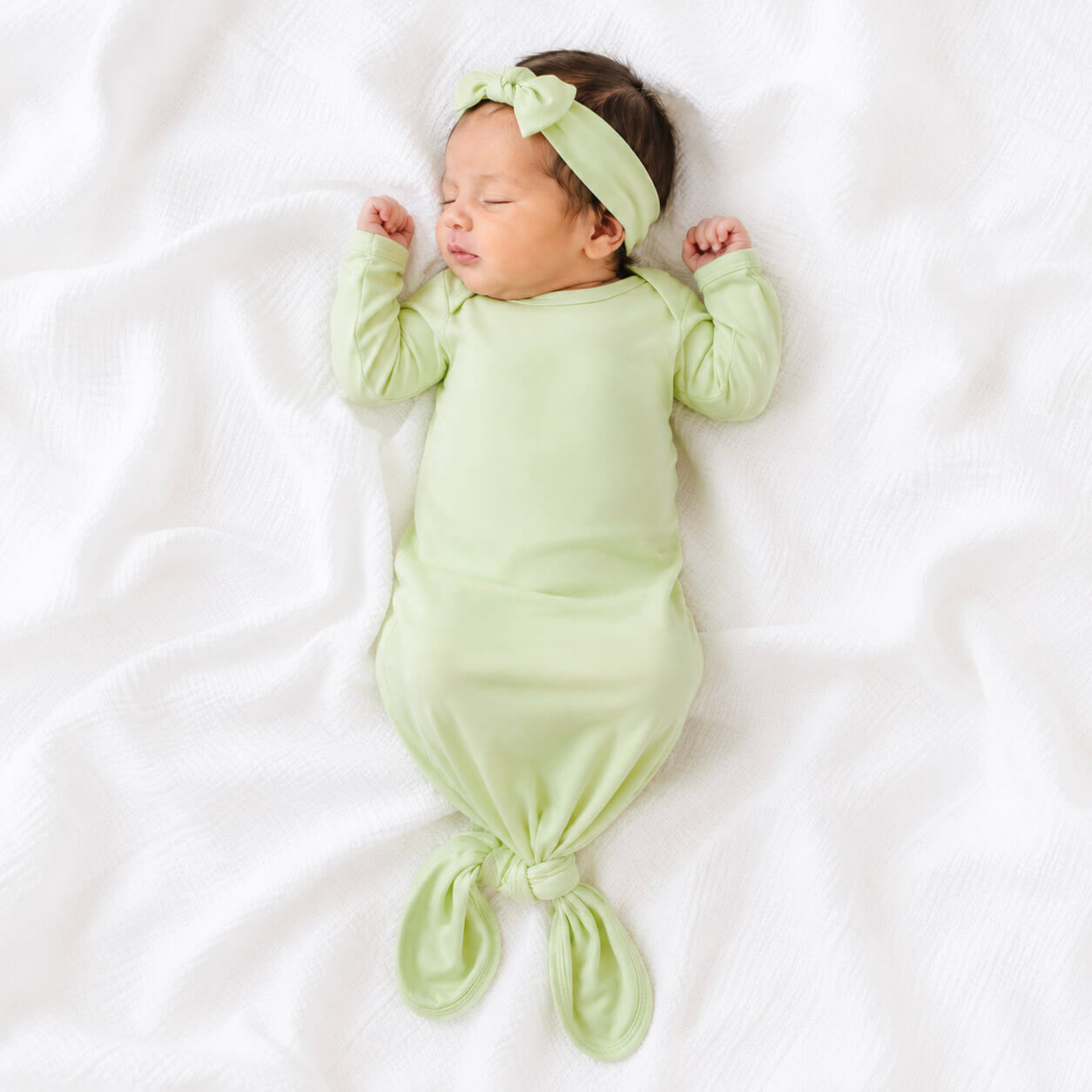 A sleeping newborn baby wearing a light green gown with a matching knotted headband, lying on a white textured blanket.