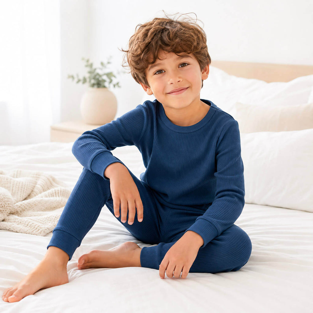 A young boy with curly brown hair sits on a white bed wearing a navy blue ribbed long-sleeve pajama set. The background shows a softly lit bedroom with white bedding, pillows, and a small potted plant on a bedside table.