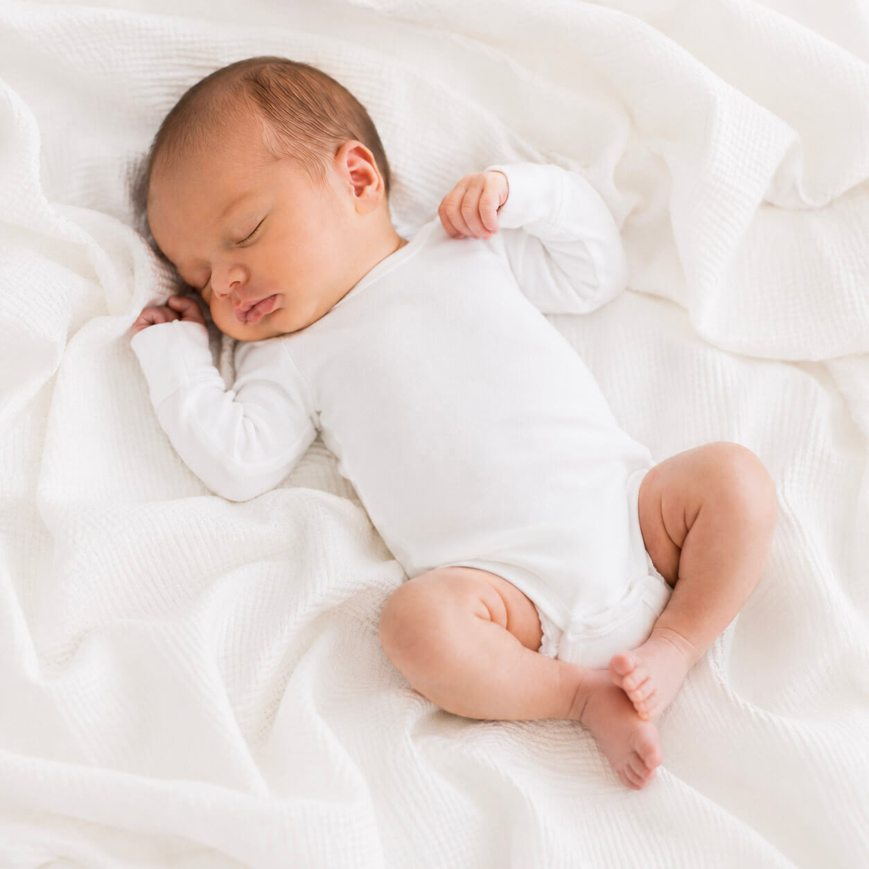 Newborn baby sleeping peacefully in a white onesie on a soft white blanket.