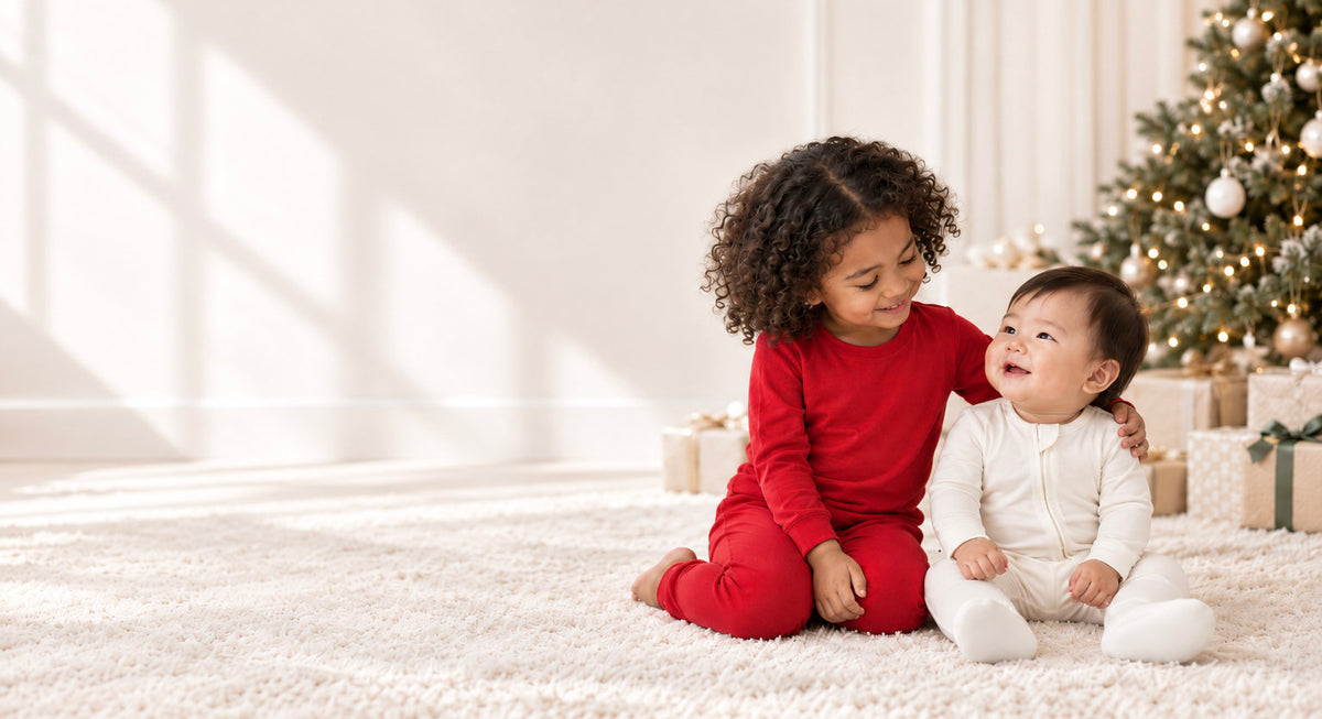 A young girl in a red long-sleeve pajama set sits on a white shag rug next to a baby in a white matching pajama set, with a decorated Christmas tree and wrapped gifts in the background.