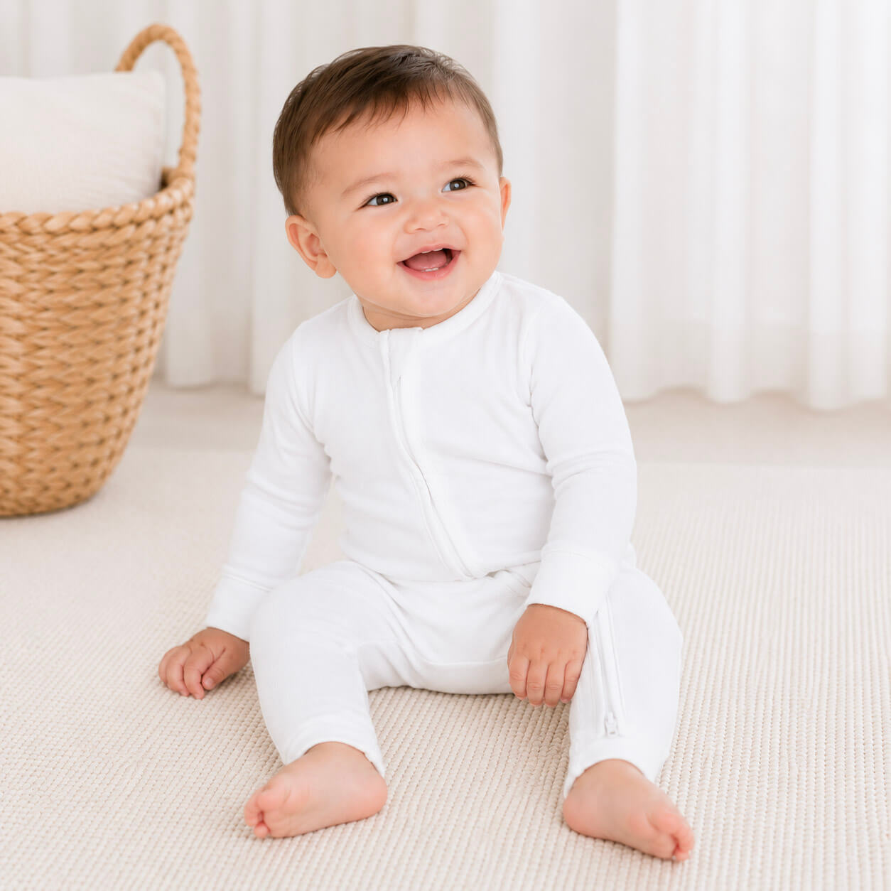 Baby wearing a white onesie sitting on a light-colored surface with a woven basket in the background.
