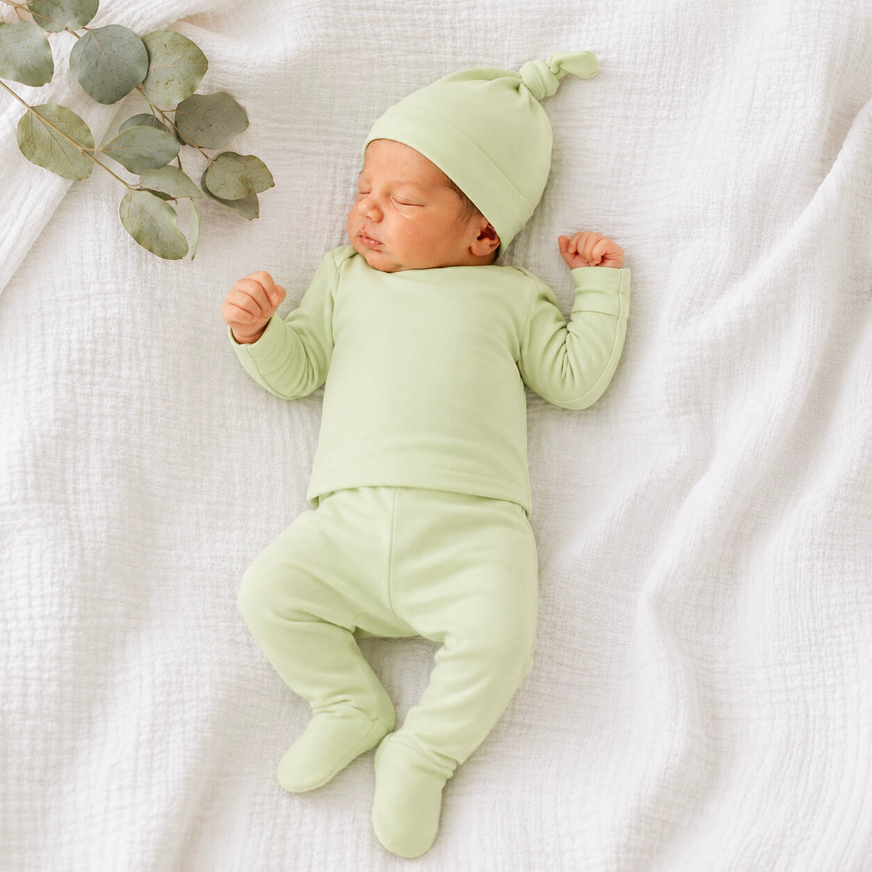 A sleeping newborn baby wearing a light sage green three-piece outfit consisting of a long-sleeve top, matching footed pants, and a knotted beanie hat, lying on a textured white muslin blanket with a eucalyptus sprig in the corner.