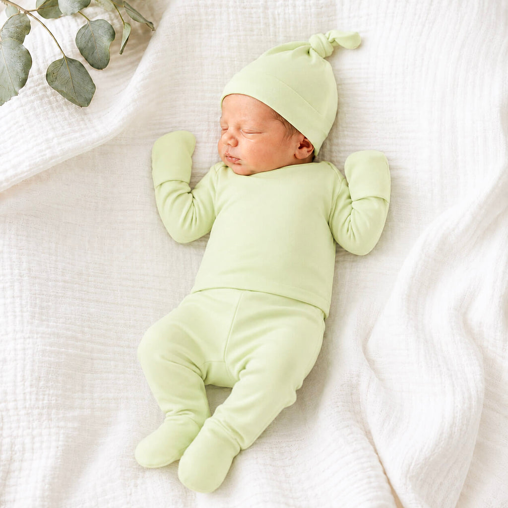 A sleeping newborn baby wearing a light sage green long-sleeve footed sleeper, matching beanie hat, and mittens, lying on a textured white muslin blanket with a eucalyptus sprig in the corner.