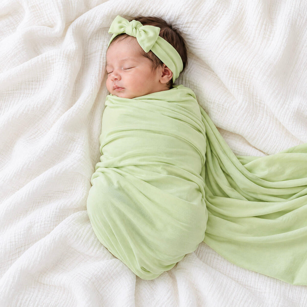 A sleeping newborn baby wrapped in a light green muslin swaddle blanket and matching bow headband, lying on a white textured muslin sheet.