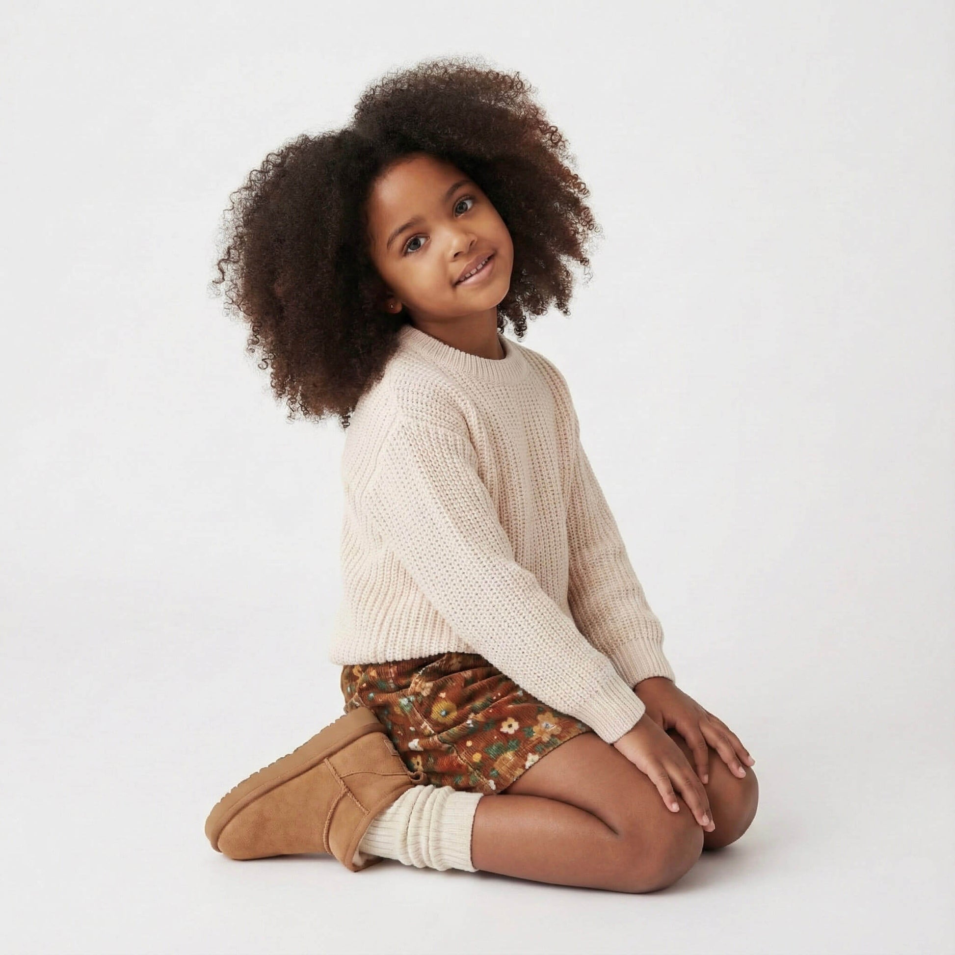 Child wearing a beige sweater and brown boots sitting on a white background