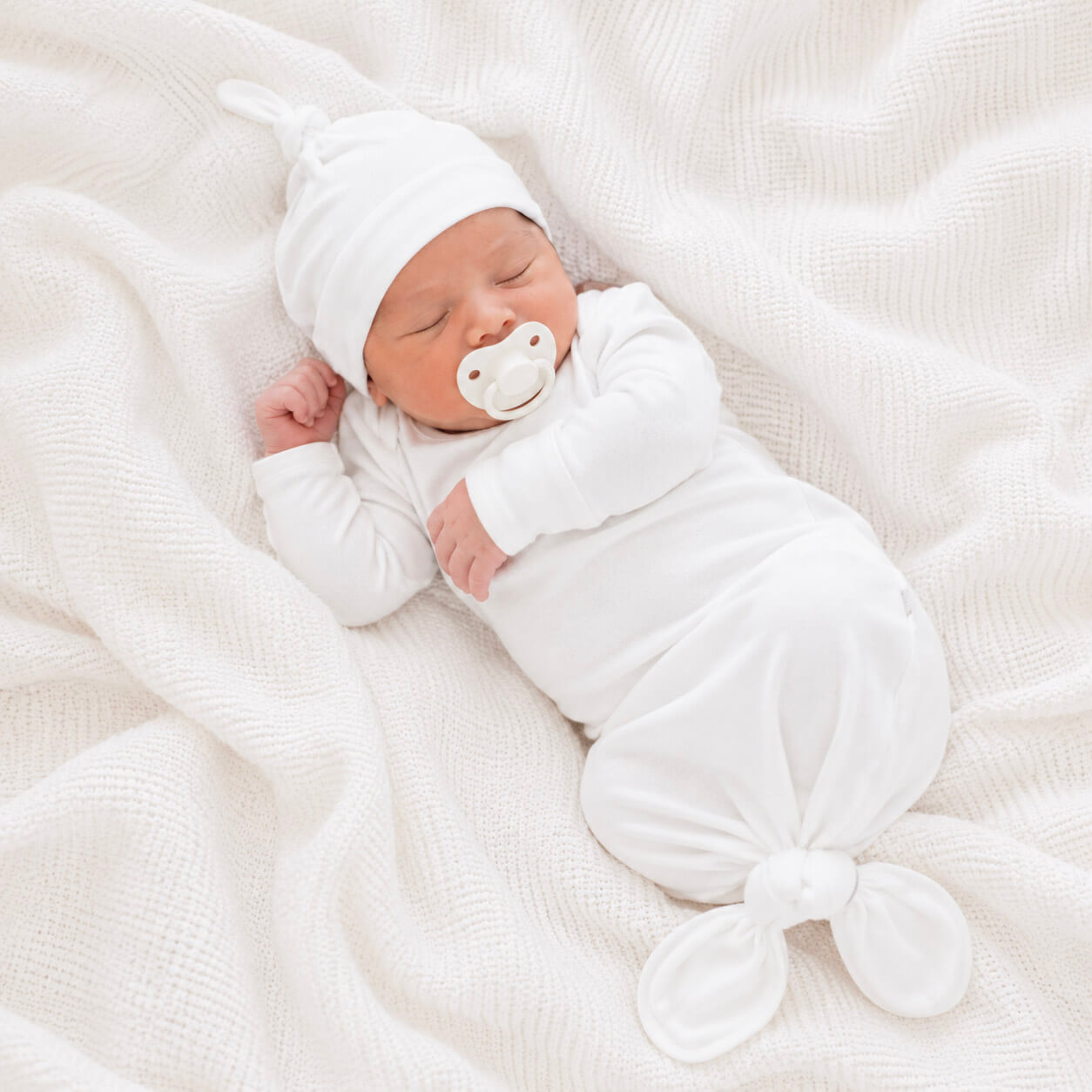 Newborn baby wrapped in white gown with a pacifier, lying on a soft white blanket.