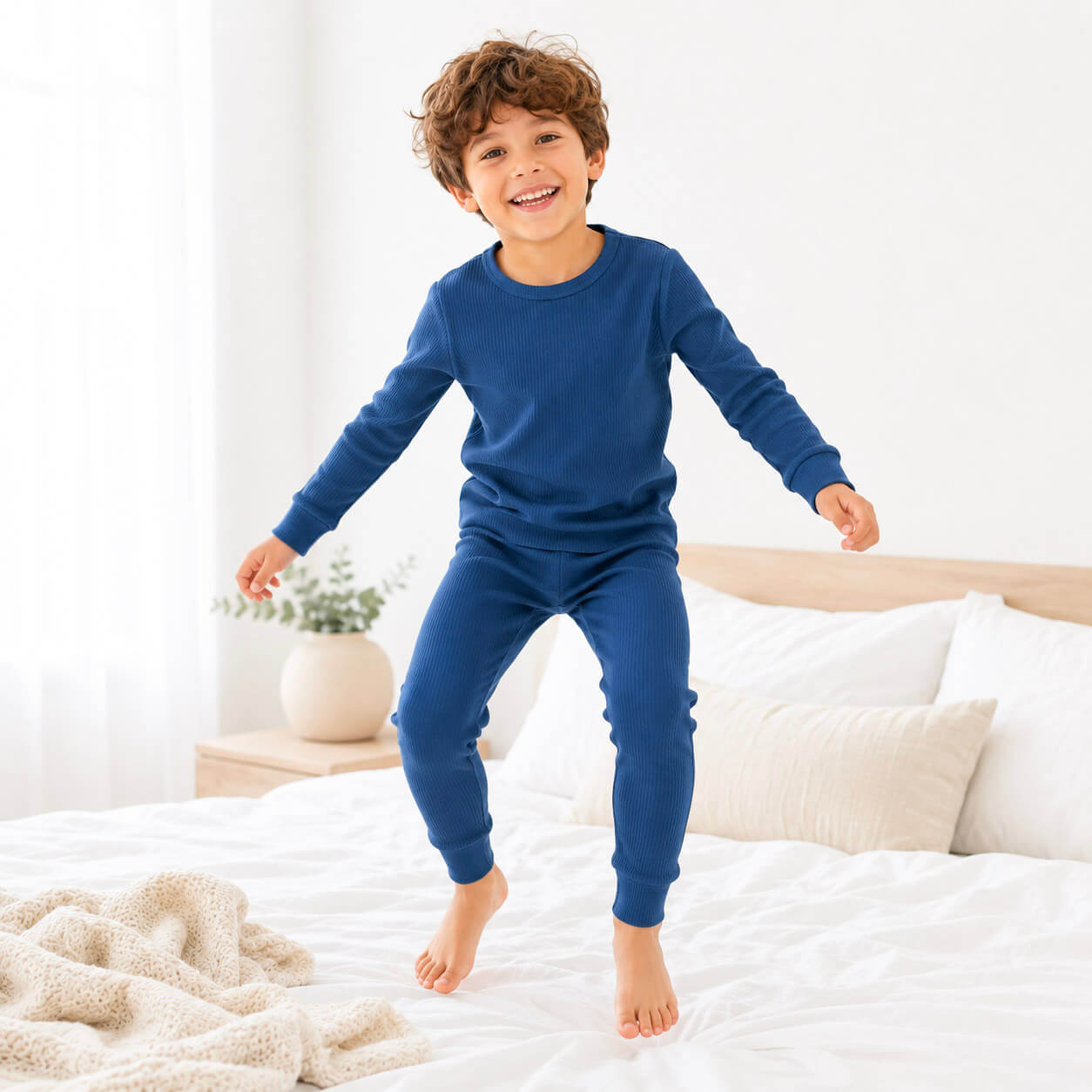 A young boy wearing a matching navy blue long-sleeve top and pants pajama set jumps on a white bed in a bright bedroom.