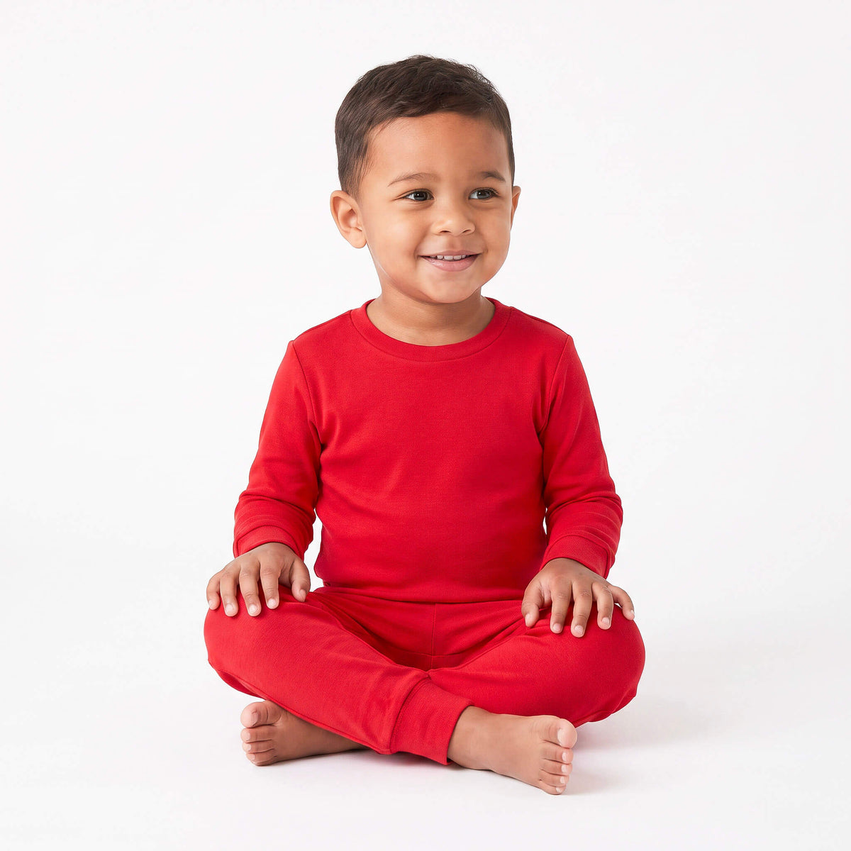 A smiling toddler boy sitting cross-legged in a bright red long-sleeve pajama set against a clean white studio background.