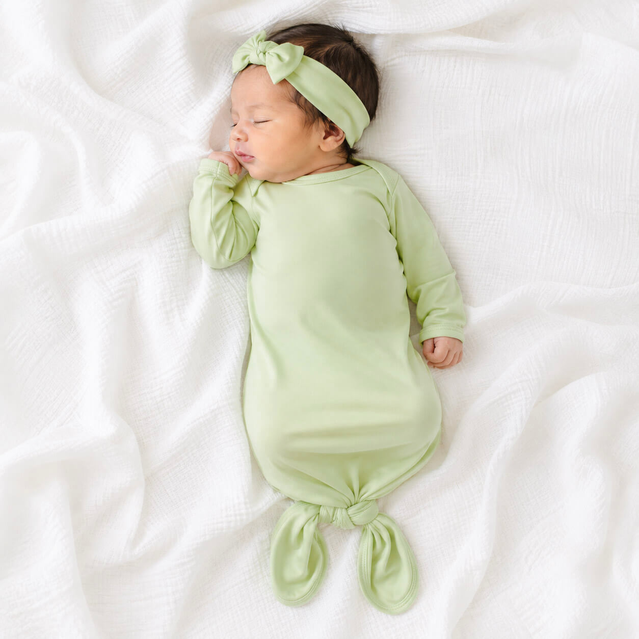 A sleeping newborn baby wearing a light green gown with a matching headband and tie at the feet, lying on a white textured blanket.