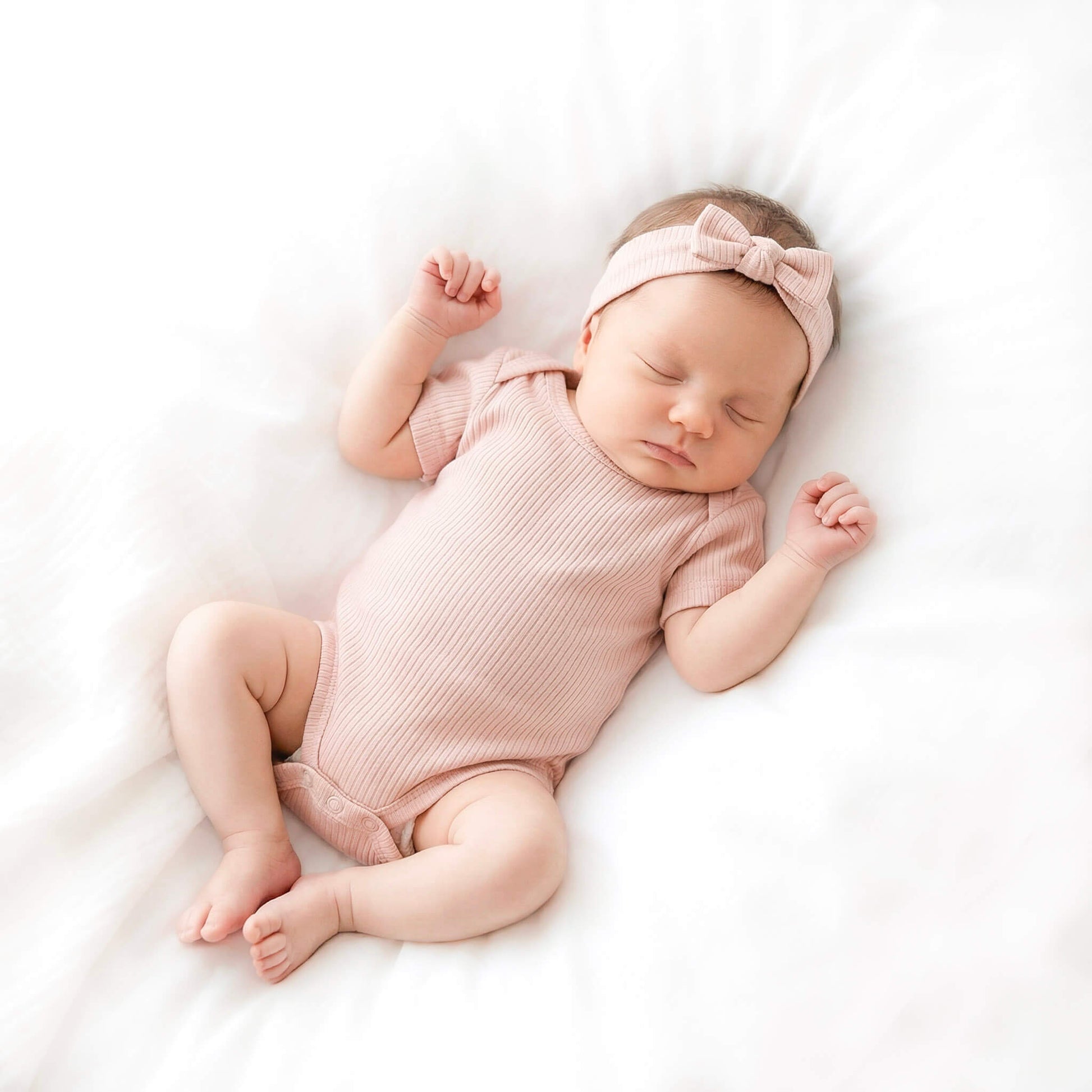Newborn baby in a pink outfit and headband sleeping on a white blanket