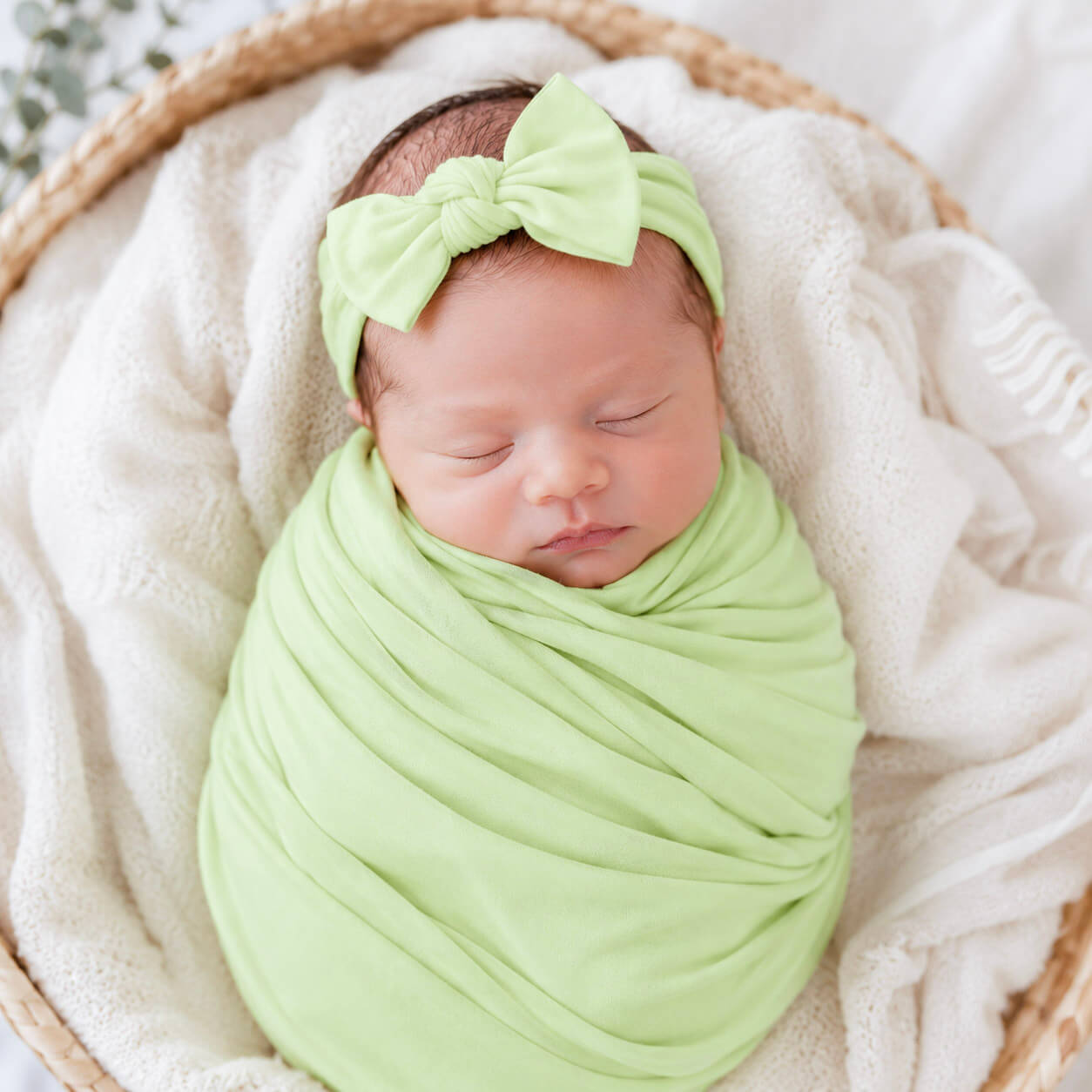 Newborn baby wrapped in a green blanket with a matching headband, lying on a white blanket.