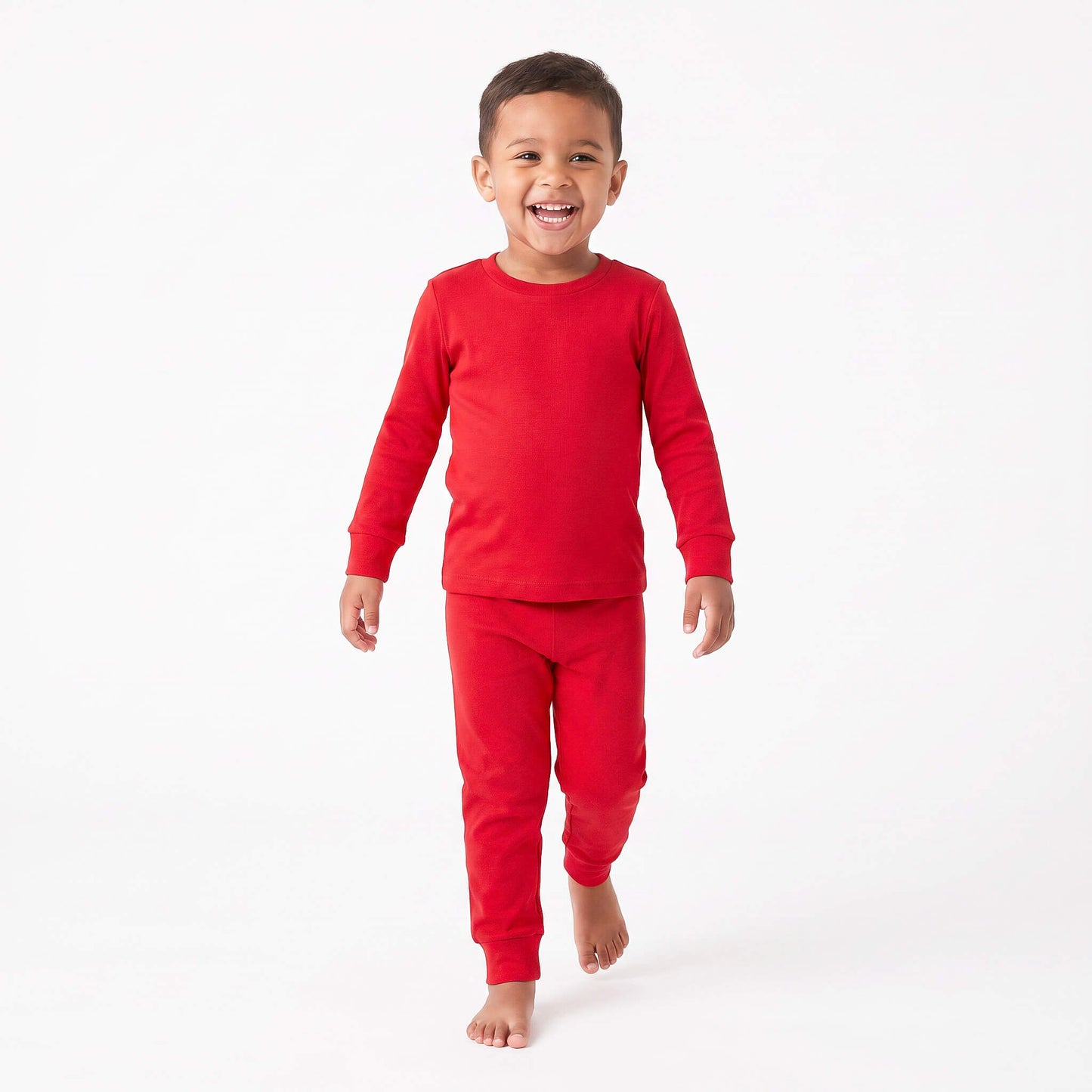 A young boy smiling and walking forward while wearing a solid red long-sleeve crewneck shirt and matching red pants, photographed against a seamless white studio background.