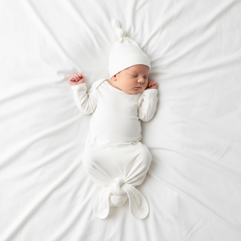 Newborn baby wrapped in a white swaddle and cap on a white bed.