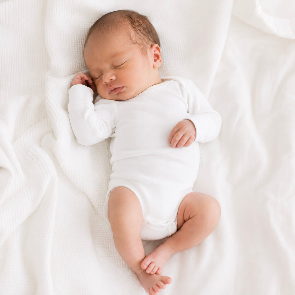 Newborn baby sleeping peacefully in a white onesie on a white blanket.