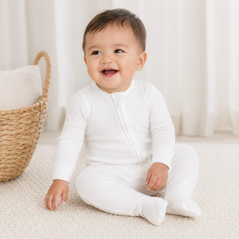Baby wearing a white onesie sitting on a light-colored surface with a woven basket in the background.